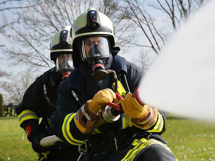 es sind zwei Mitarbeiter der Feuerwehr in Arbeitskleidung abgebildet. Sie betätigen einen Wasserschlauch. 