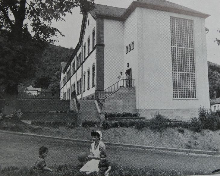 Ein Schwarz-Weiß-Foto von der jugendpsychiatrischen Abteilung und Beobachtungsstation für ruhige männliche Patienten der damaligen Pfälzischen Nervenklinik Landeck in den 50er Jahren. Im Hintergrund das Gebäude, im Vordergrund eine Krankenschwester mit 2 Jungen beim Ball spielen.