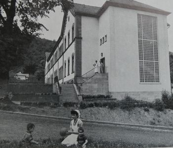 Ein Schwarz-Weiß-Foto von der jugendpsychiatrischen Abteilung und Beobachtungsstation für ruhige männliche Patienten der damaligen Pfälzischen Nervenklinik Landeck in den 50er Jahren. Im Hintergrund das Gebäude, im Vordergrund eine Krankenschwester mit 2 Jungen beim Ball spielen.