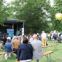 Eine Bühne ist auf einer Wiese zu sehen, davor Menschen auf Festbänken.