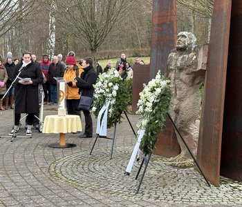 Frauen berichten in einer Andacht über das Schicksal von Maria Z. und anderen Patient*innen, im Vordergrund die Skulptur auf der Gedenkstätte mit den Kränzen, im Hintergrund Besucher*innen.