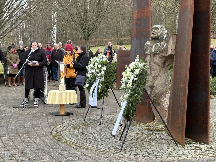 Frauen berichten in einer Andacht über das Schicksal von Maria Z. und anderen Patient*innen, im Vordergrund die Skulptur auf der Gedenkstätte mit den Kränzen, im Hintergrund Besucher*innen.