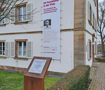 Das Bild zeigt das Alleehaus mit einem Werbebanner für die Ausstellung und einer Gedenkstele, die davor steht.