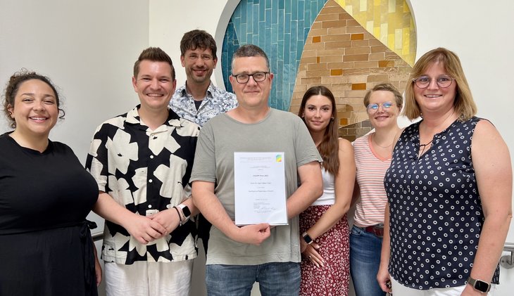2025-07-07_Team_Prävent_plus_mit_Urkunde Auf dem Bild sind 7 Mitarbeitende des Pfalzklinikums zu sehen, die stolz die Urkunde zum gewonnen Preis in die Kamera halten.