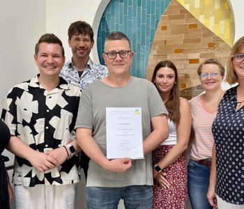 2025-07-07_Team_Prävent_plus_mit_Urkunde Auf dem Bild sind 7 Mitarbeitende des Pfalzklinikums zu sehen, die stolz die Urkunde zum gewonnen Preis in die Kamera halten.