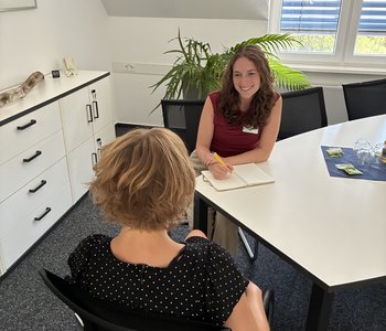 Lara Heß, eine junge Frau mit langen dunklen Haaren, sitzt mit einem Notizbuch am Tisch und berät eine Klientin.