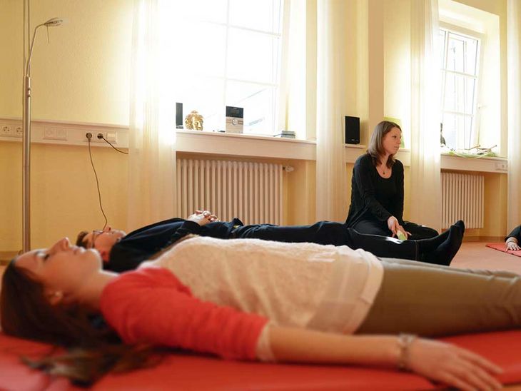 Therapie im Pfalzklinikum besteht auch aus bewusster Entspannung. Auf diesem Bild sieht man mehrere Patientinnen und Patienten, die auf Matten auf einem weichen Boden liegen. Zwischen ihnen sitzt eine Therapeutin im Schneidersitz und leitet die Entspannungsübung.