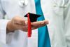 Symbolbild: Mediziner im weißen Kittel hält Mini-Doktorandenhut in der Hand 
