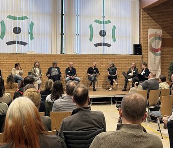 Podiumsdiskussion auf dem Symposium. 9 Menschen sitzen im Halbkreis in einem Gespräch auf einer Bühne.