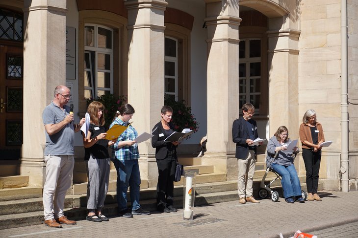 Das Foto zeigt 8 Mitglieder des Gedenkausschusses mit Mappen und Blättern vor dem Hauptgebäude beim Vorlesen. Davor steht die brennende Gedenkkerze.