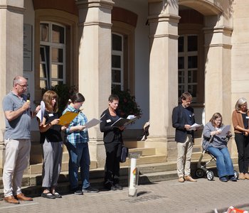 Das Foto zeigt 8 Mitglieder des Gedenkausschusses mit Mappen und Blättern vor dem Hauptgebäude beim Vorlesen. Davor steht die brennende Gedenkkerze.