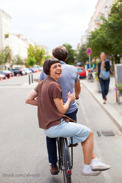 Symbolbild: Ein Fahrrad fährt auf der Straße, eine Frau sitzt auf dem Gepäckträger und winkt lachend in die Kamera
