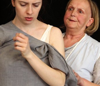 Eine ältere Frau steht hinter einer jüngeren Frau und zieht ihr das Kleid aus - Szene aus dem Theaterstück "Rosa B. - beinahe vergessen"
