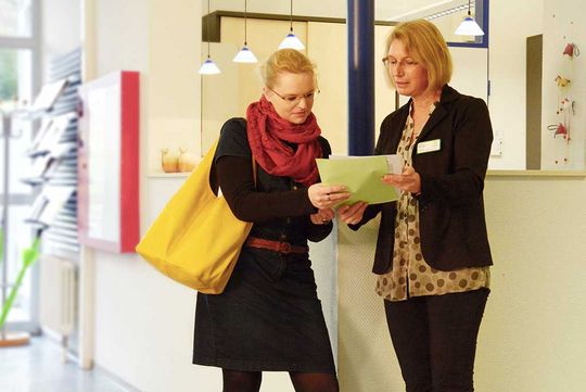 zwei Frauen stehen vor der Aufnahme und schauen auf einen Zettel welcher eine der Frauen in der Hand hält