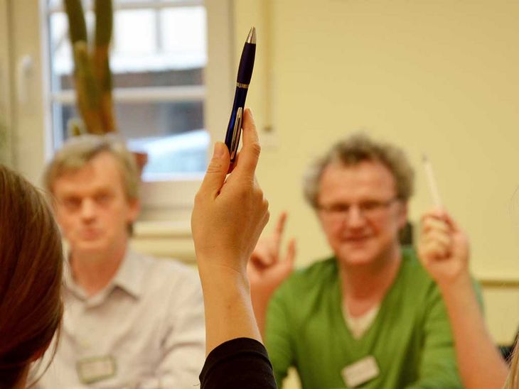 drei Personen sind in einer Gesprächssituation. Eine Person meldet sich mit einem Stift in der Hand 