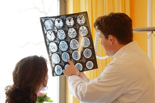 Ein Arzt ist im Gespräch mit einer Patientin in einem freundlich gestalteten Zimmer der Klinik für Neurologie. Er zeigt ihr CT oder Röntgenaufnahmen und erklärt ihr die Befunde der Untersuchung. Die Patientin ist nur von hinten zu sehen, der Arzt ebenfalls.