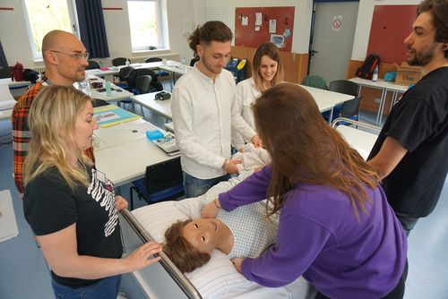 Schüler*innen und eine Lehrerin üben mithilfe einer Puppe an einem Krankenbett.