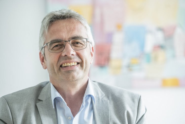 Das Bild zeigt Professor Markus Steffens. Er hat graues Haar, trägt eine Brille und einen grauen Anzug. Im Hintergrund ist ein buntes Bild an der Wand zu sehen.