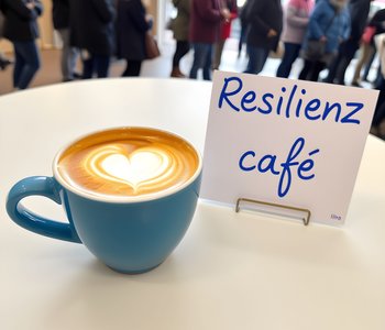 Das Bild zeigt eine Tasse mit Kaffee, ein Schild mit dem Schriftzug "Resilienzcafé" und im Hintergrund eine Gruppe Menschen
