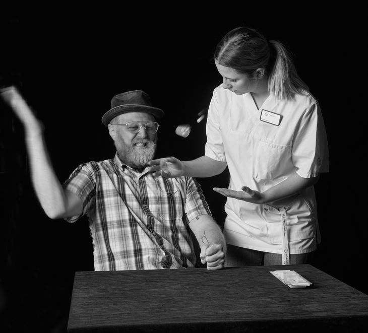 Ein alter Mann wirft mit Tabletten, eine Pflegekraft versucht, ihn zu beruhigen.