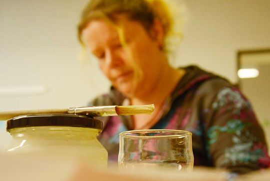 Im Vordergrund des Bildes sieht man einen Malpinsel und im Hintergrund ist eine Frau abgebildet