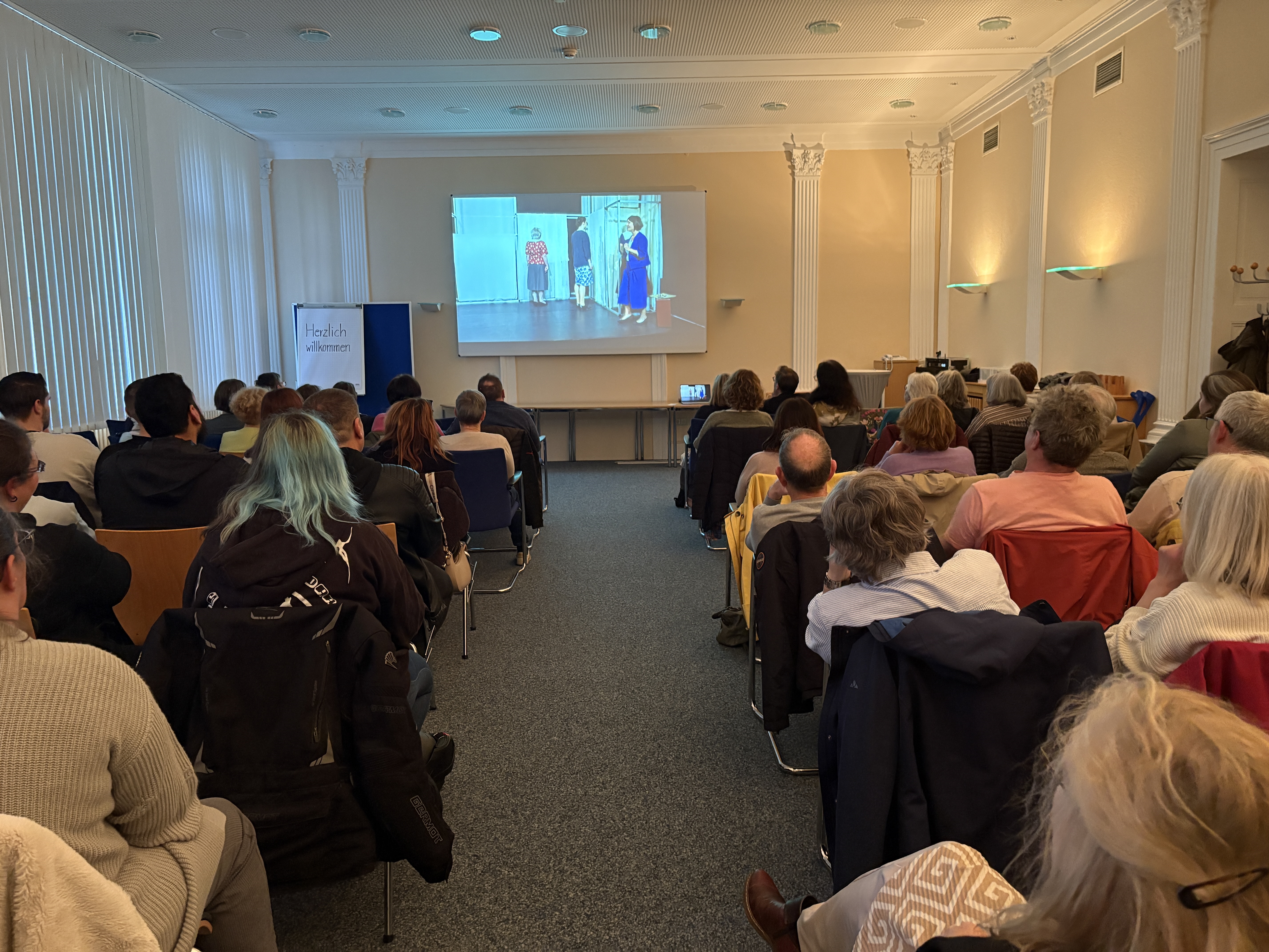 Das Bild zeigt den Konferenzraum mit dem Publikum und die Leinwand mit dem Video des Theaterstücks.