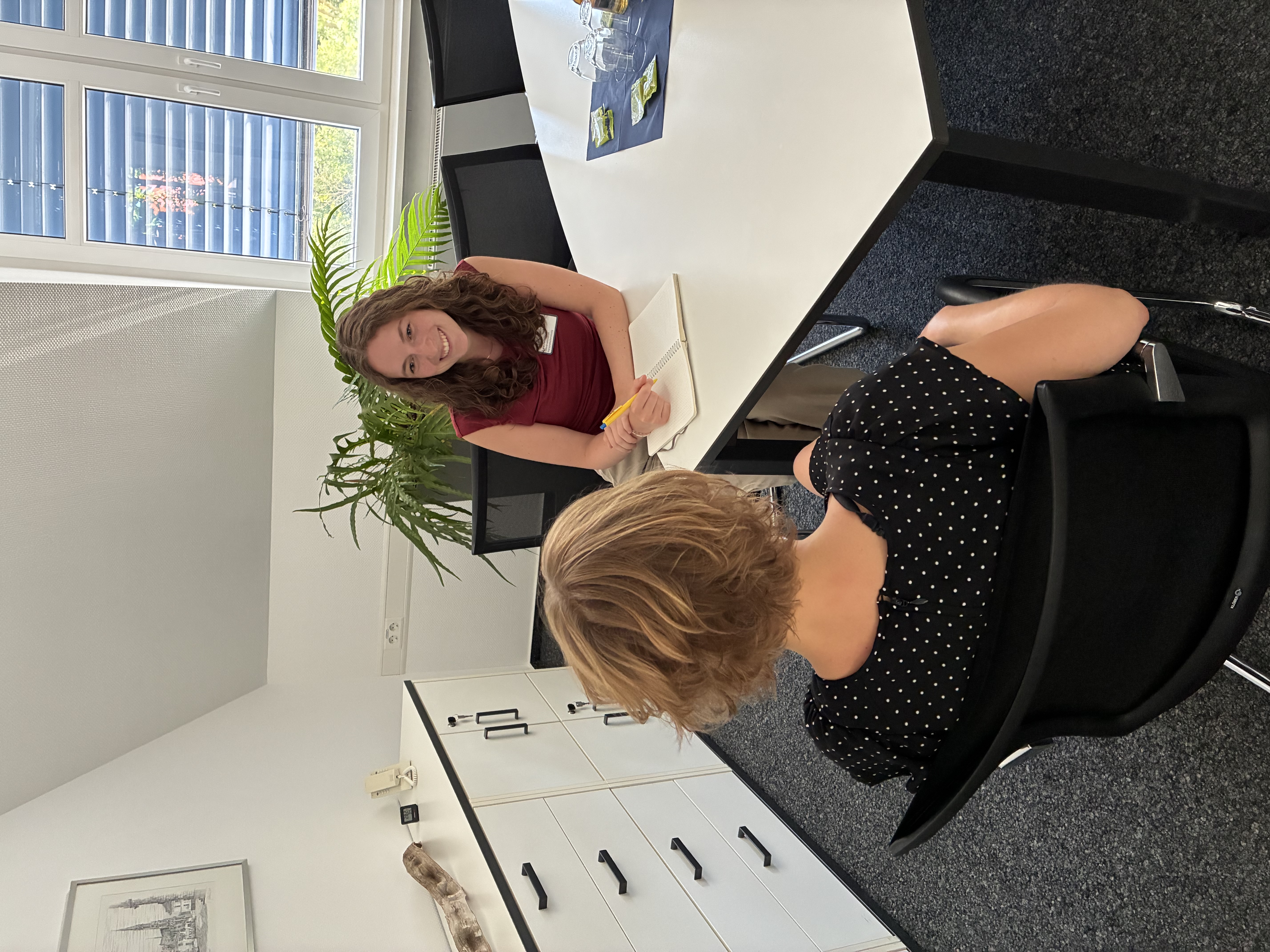 Lara Heß, eine junge Frau mit langen dunklen Haaren, sitzt mit einem Notizbuch am Tisch und berät eine Klientin, die vor ihr sitzt.