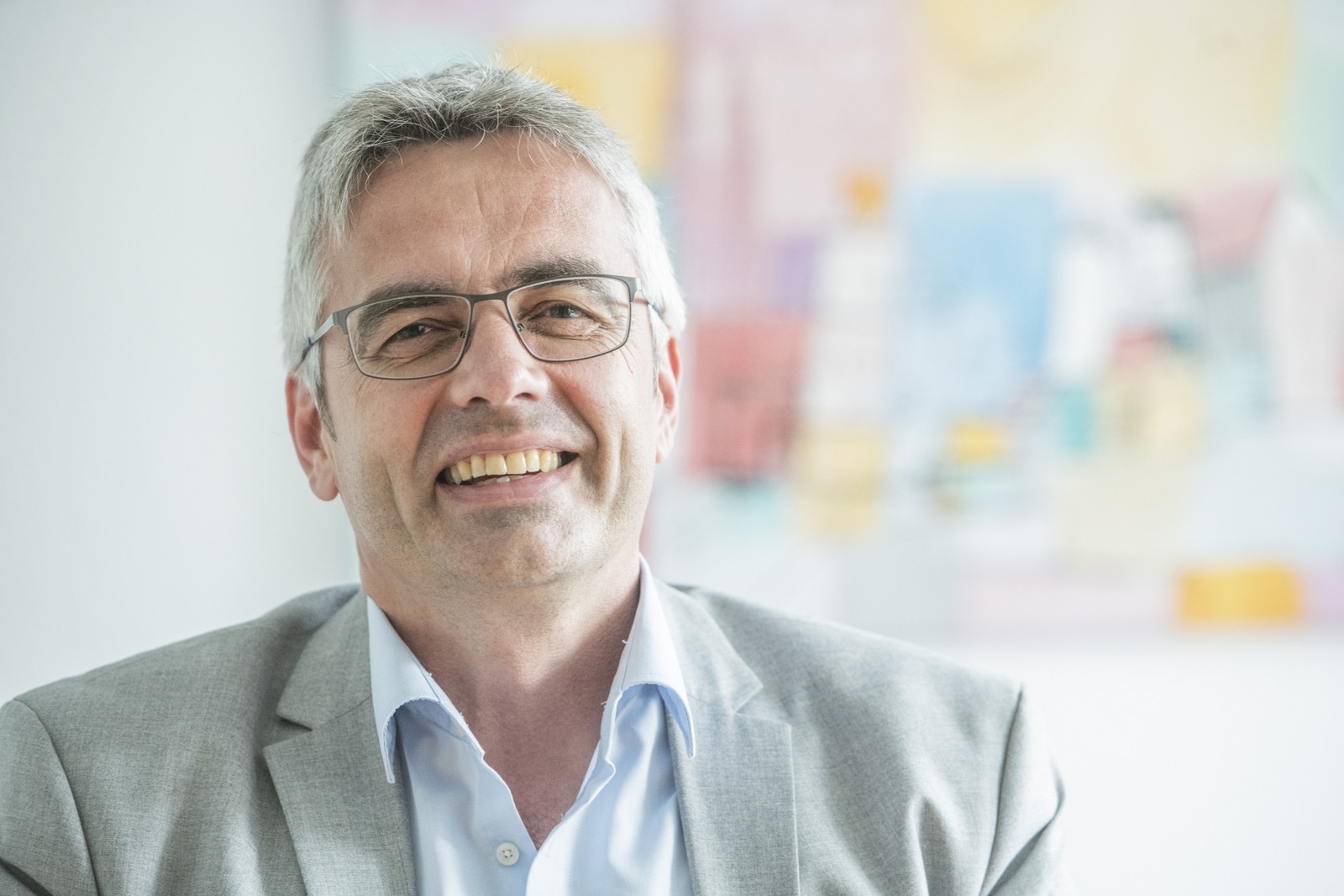 Das Bild zeigt Professor Markus Steffens. Er hat graues Haar, trägt einen grauen Anzug und eine Brille. Im Hintergrund ist ein buntes Bild an der Wand zu sehen.