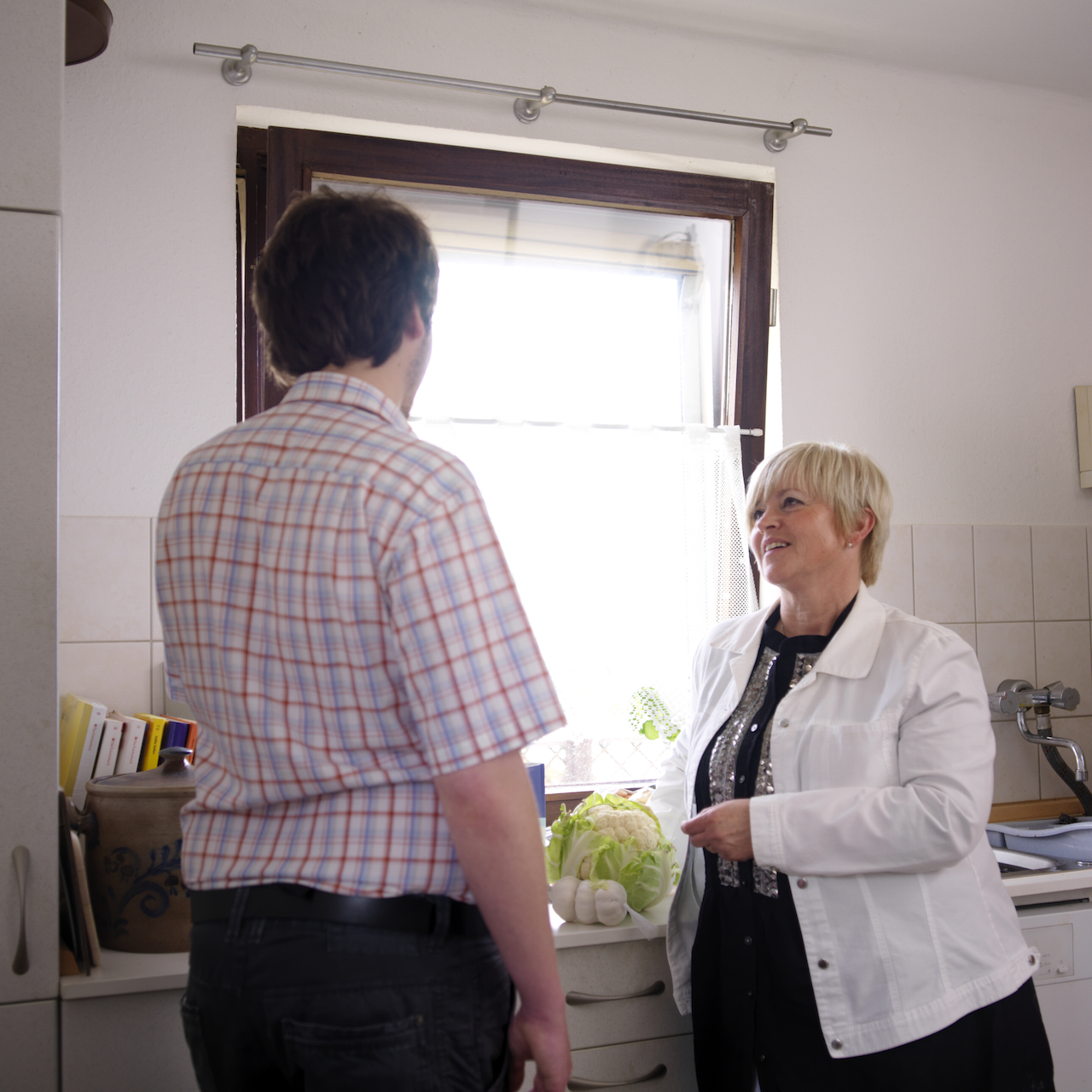 Eine Frau steht einem Mann gegenüber. Sie steht mit ihm in einer Küche und bespricht mit ihm, welche Lebensmittel er benötigt. Sie ist Mitarbeiterin der ambulanten psychiatrischen Pflege und Betreuung des Pfalzklinikums. Im Hintergrund sind ein Kopf Salat, Knoblauchzehen in einem Säckchen sowie die Spüle zu sehen. 